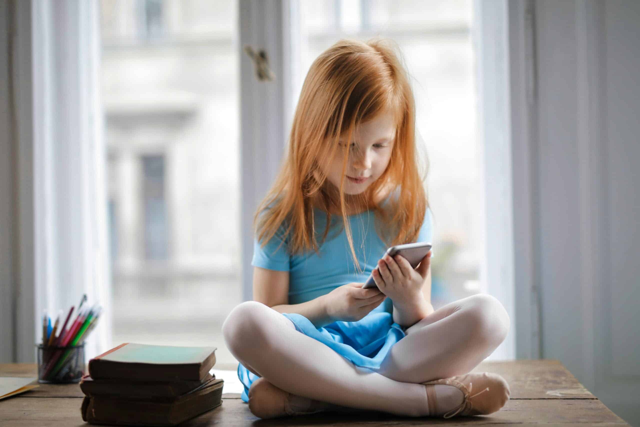 A small ginger girl sitting on a table, using a smartphone.