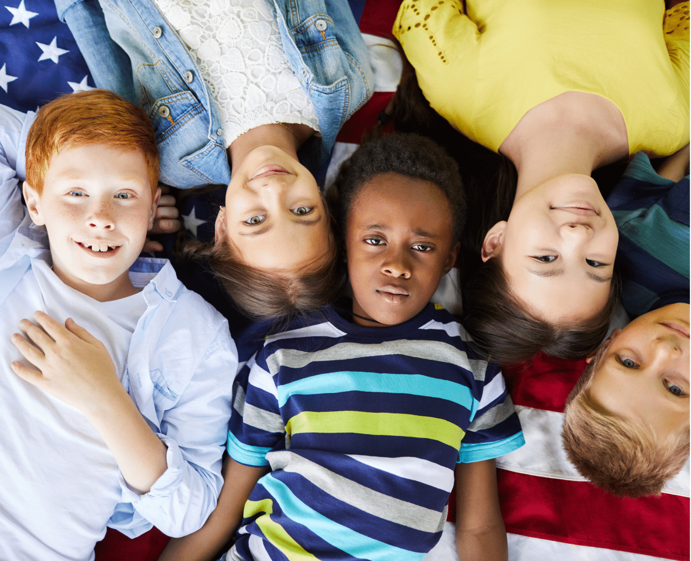 A group of five children lying down on an American flag, their heads close together in a circle. They appear to be from diverse backgrounds, reflecting a sense of unity and inclusion. The children are looking up at the camera, with expressions ranging from neutral to slightly smiling. The group consists of boys and girls, each dressed in casual clothing with a colourful variety of stripes, denim, and solid colours.