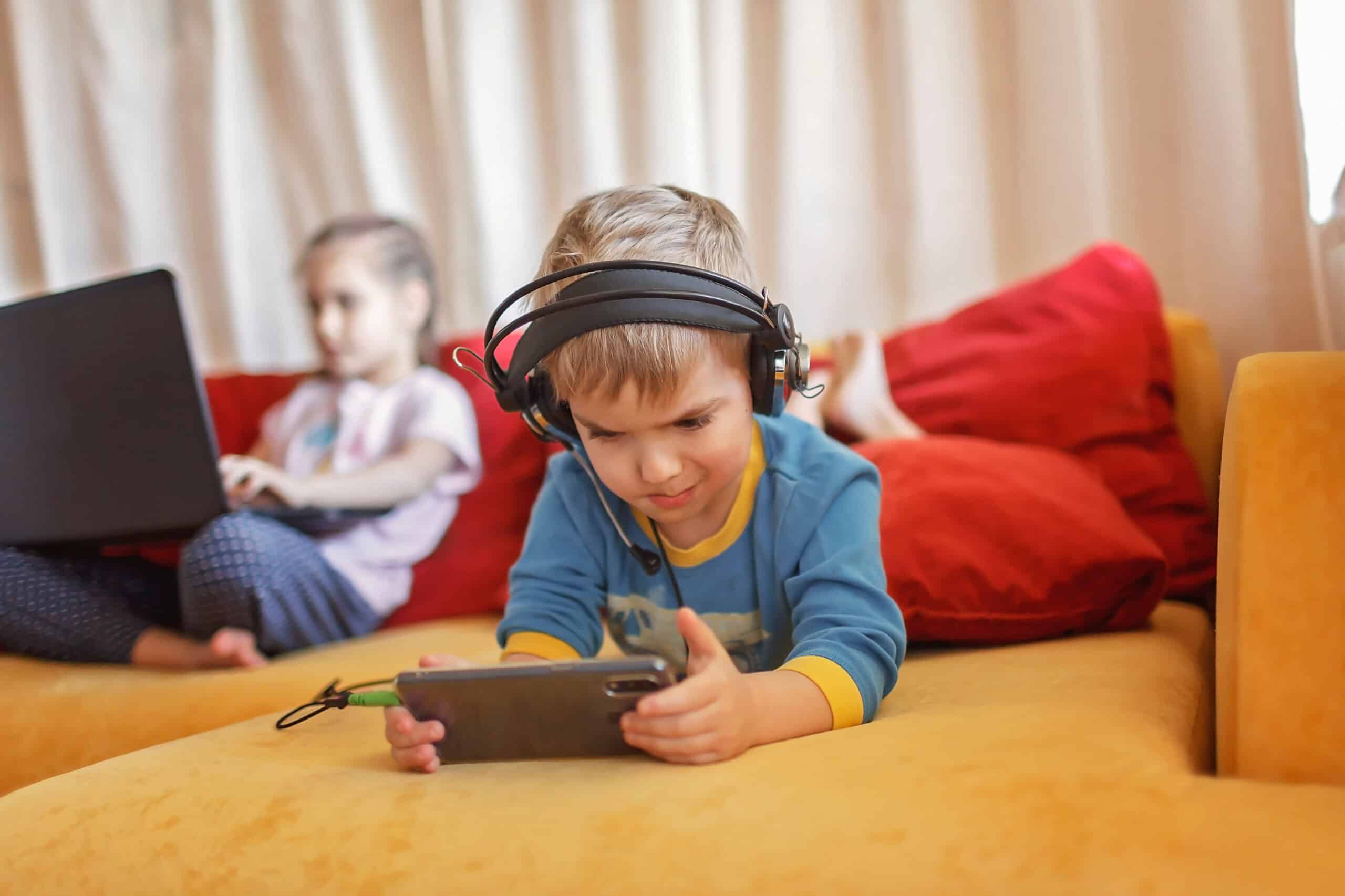 A young boy is lying down on a yellow sofa. He is lying on his front with his space position towards the camera. He is wearing headphones and looking intently at a phone. A young girl is in the background, on the sofa, on a laptop, but out of focus.