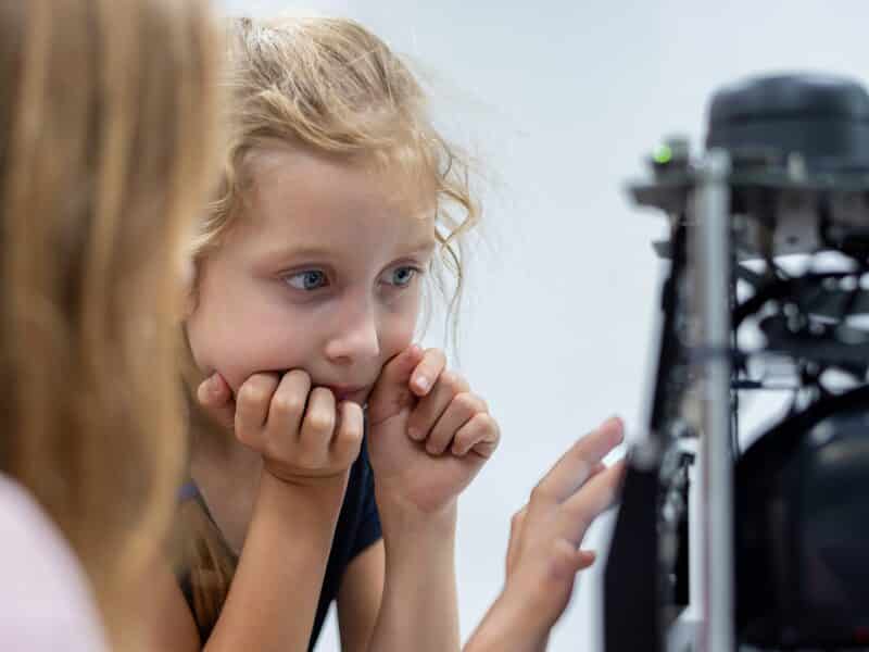 A young blonde girl enamoured by a machine learning robot. A woman, with her back to her camera and blurry, is taping the screen.