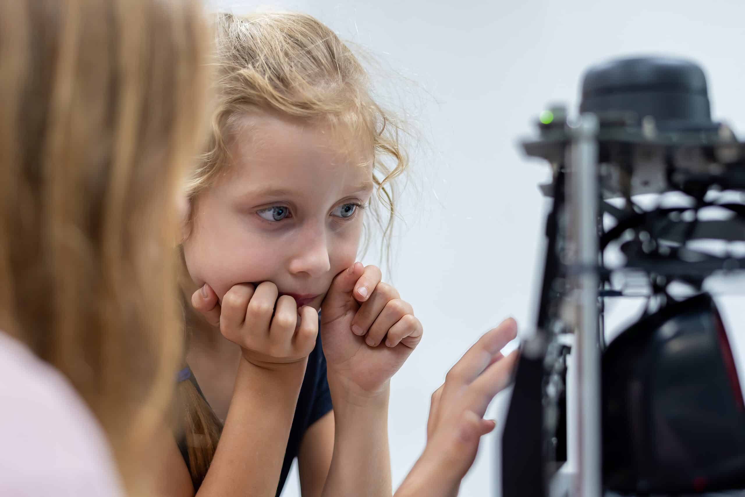 A young blonde girl enamoured by a machine learning robot. A woman, with her back to her camera and blurry, is taping the screen.