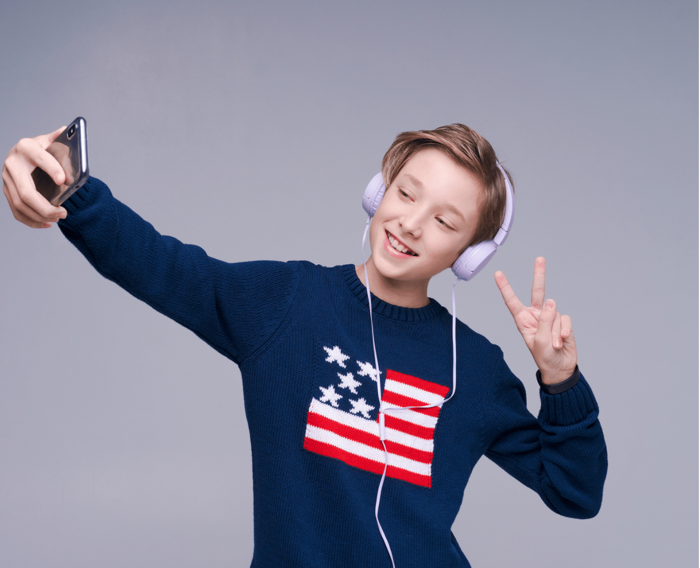 A young boy taking a selfie with a smartphone while wearing light purple headphones. He is smiling and holding up a peace sign with his other hand. The boy is wearing a dark blue sweater with an American flag design on the front.