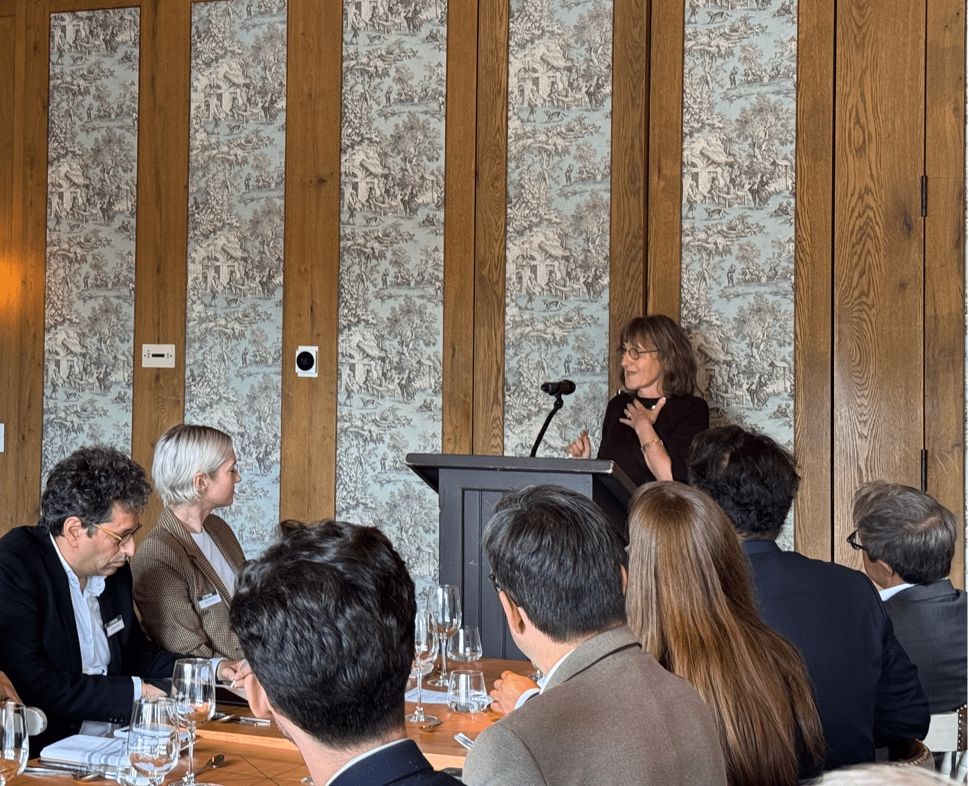 The image shows Baroness Beeban Kidron speaking at a podium during a conference in Toronto titled "ATTENTION: THE KIDS ARE ALL RIGHT." She is addressing a seated audience in a formal dining setting. The room features ornate, wood-paneled walls with detailed wallpaper in a pastoral design. The audience members, some of whom are listening attentively, are seated around tables set with glassware. 