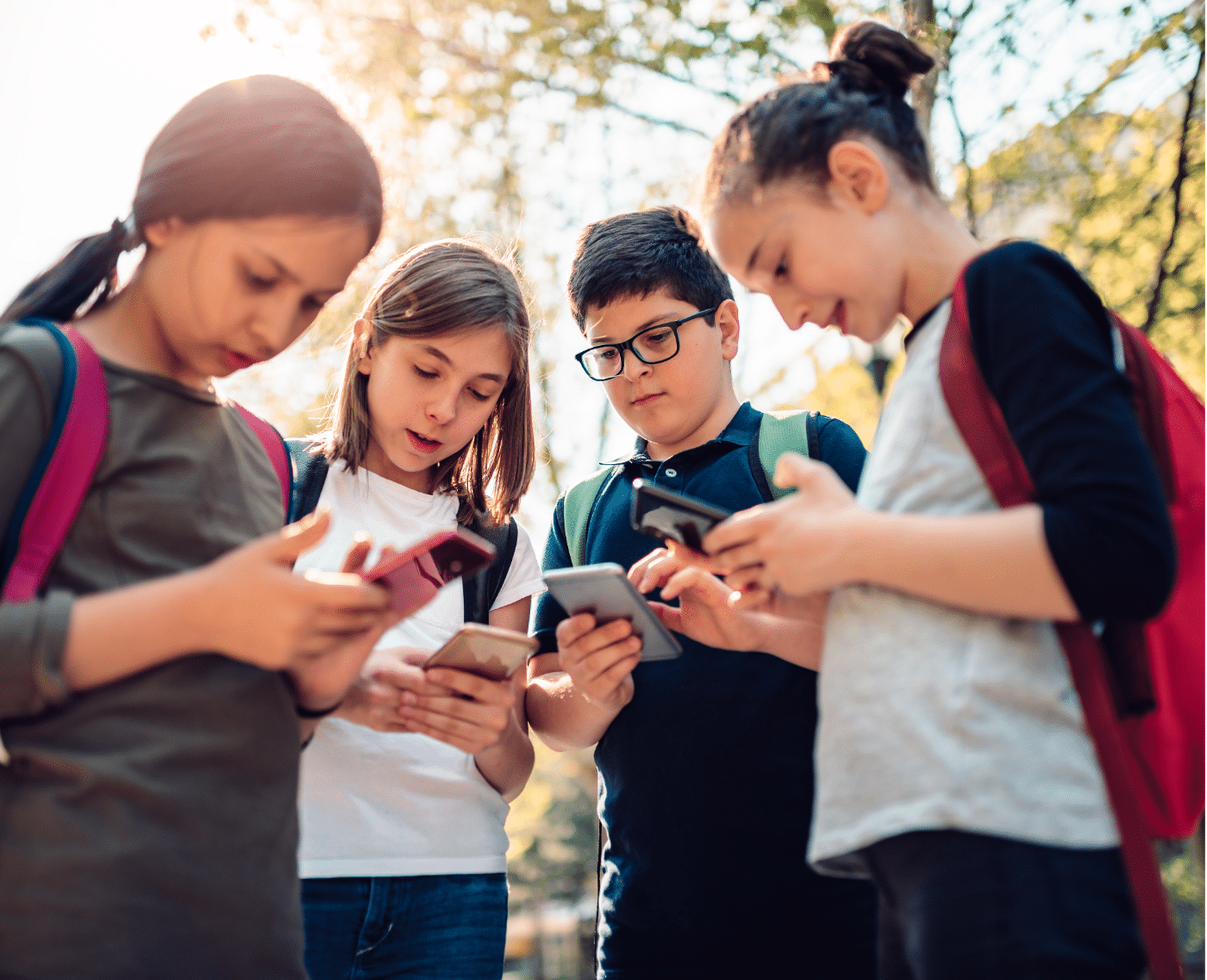 A group of four children standing close together, each focused on their smartphones. They appear to be pre-teens or early adolescents, dressed casually with backpacks, suggesting they might be on their way to or from school. The children are absorbed in their devices, seemingly engaged in a shared activity, though each one is holding and looking at their own phone. 