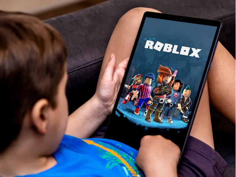 A wider shot showing the same child sitting on a couch, holding the tablet while playing Roblox. The screen features colorful characters from the game, and the child is wearing a blue T-shirt. The entire tablet and part of the child’s seated posture are visible. This is the full version of the close-up shown above.