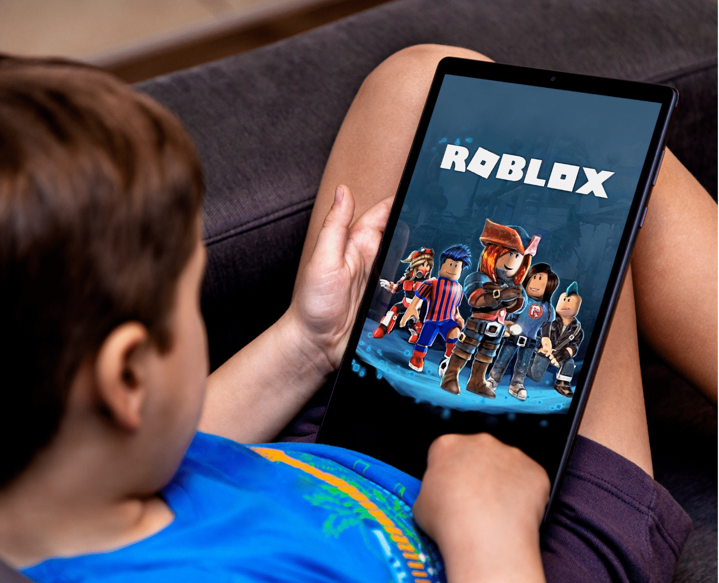 A wider shot showing the same child sitting on a couch, holding the tablet while playing Roblox. The screen features colorful characters from the game, and the child is wearing a blue T-shirt. The entire tablet and part of the child’s seated posture are visible. This is the full version of the close-up shown above.