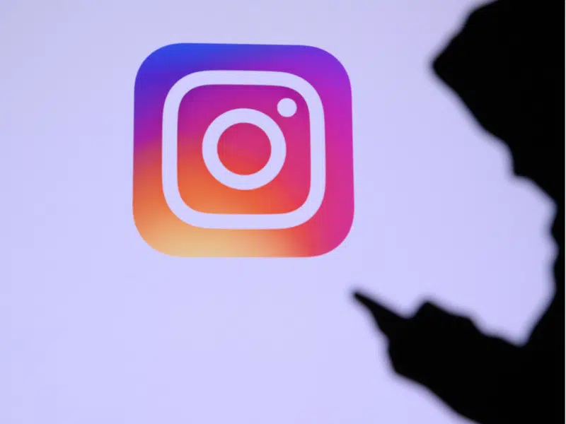 A wider shot showing the large Instagram logo on a screen. In the foreground, there is a silhouette of a person holding and using a smartphone, with the focus primarily on the logo. This is the full version of the close-up shown above.