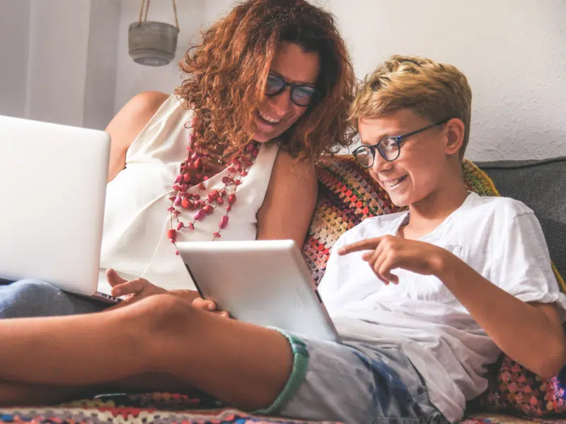 A middle-aged woman with red curly hair is sitting on the sofa with a laptop on her lap next to a blonde boy wearing rounded glasses on a tablet. Both subjects are looking at the boy's tablet and smiling.