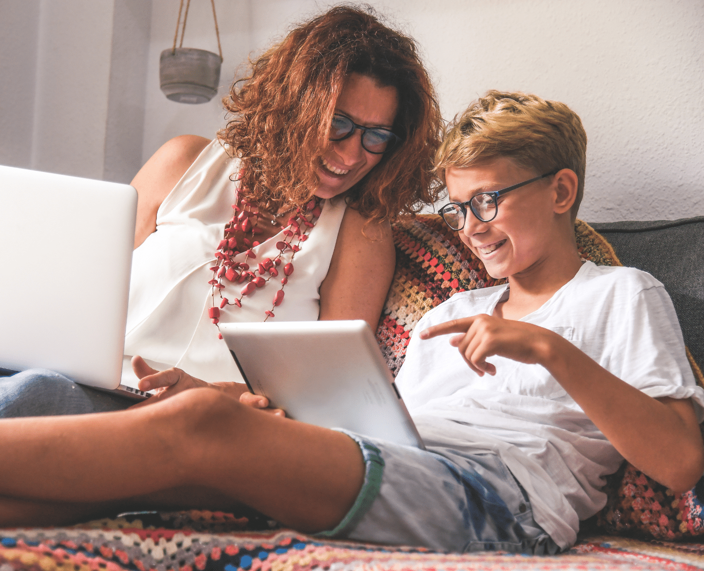 A middle-aged woman with red curly hair is sitting on the sofa with a laptop on her lap next to a blonde boy wearing rounded glasses on a tablet. Both subjects are looking at the boy's tablet and smiling. 