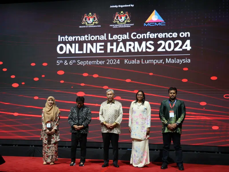 Two women and three men appeared on a red-carpeted stage. They are standing in front of a graphic that consists of red light beams being intersected by red circles. The graphic includes the words 'International Legal Conference on Online Harms 2024'.