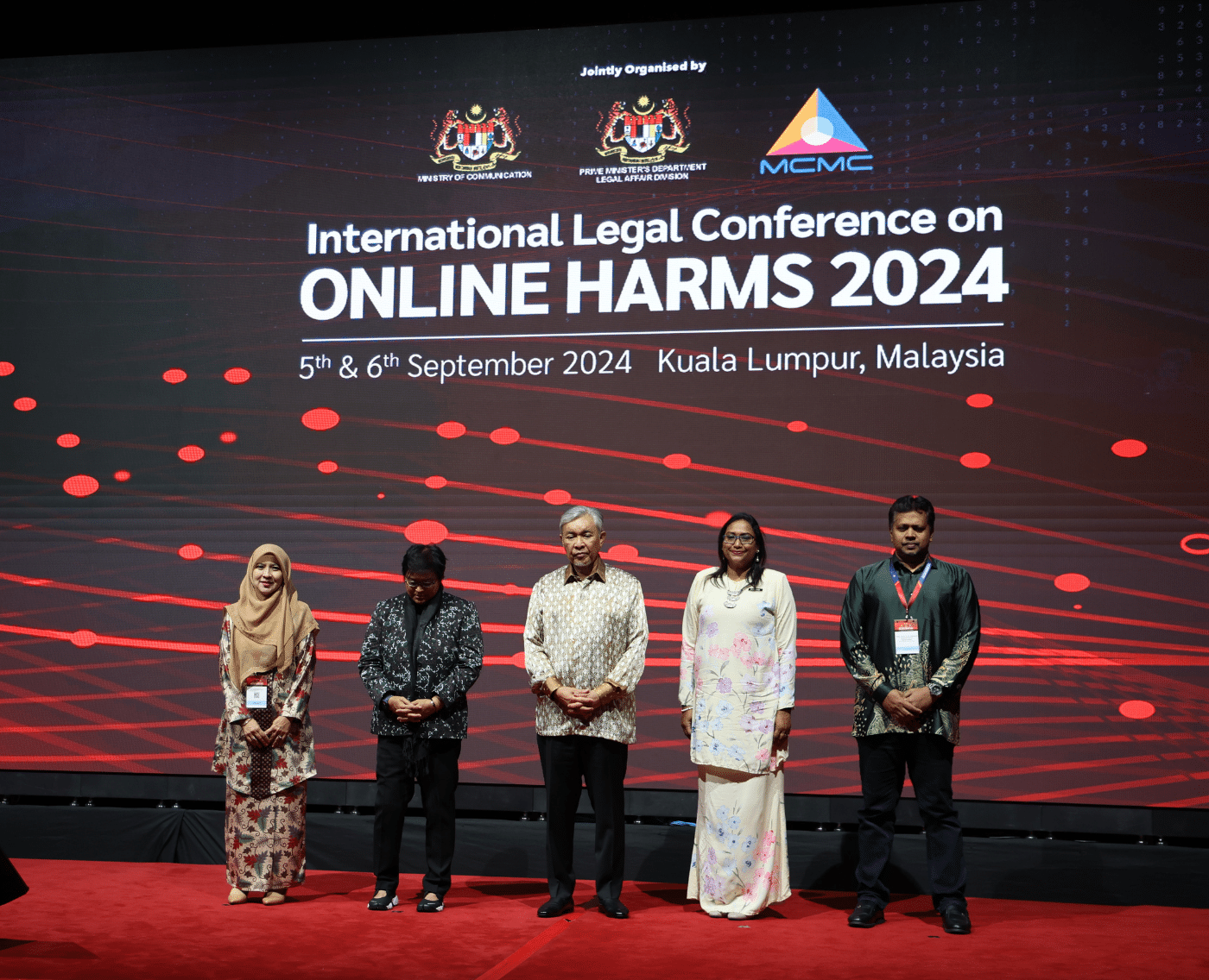 Two women and three men appeared on a red-carpeted stage. They are standing in front of a graphic that consists of red light beams being intersected by red circles. The graphic includes the words 'International Legal Conference on Online Harms 2024'. 