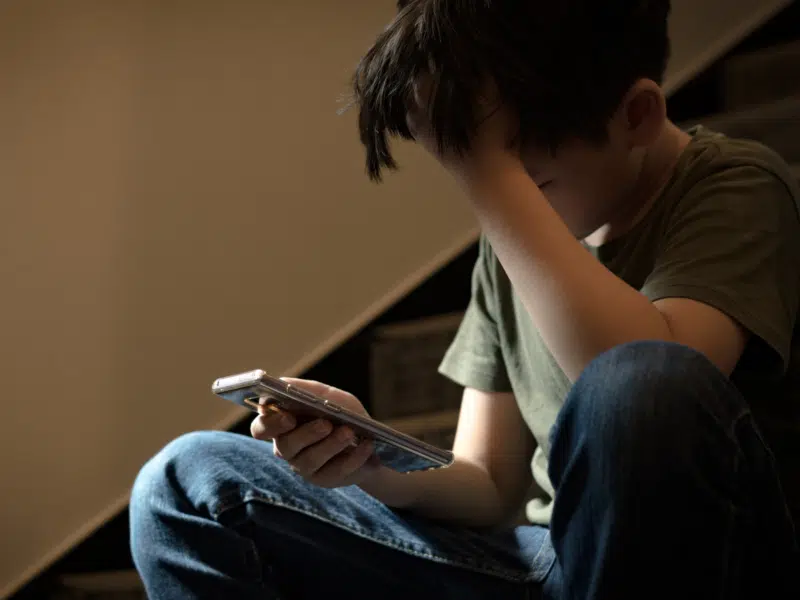 A young boy sat on a staircase. He is holding a phone in his right hand and holding his head with his left hand. He looks dejected.