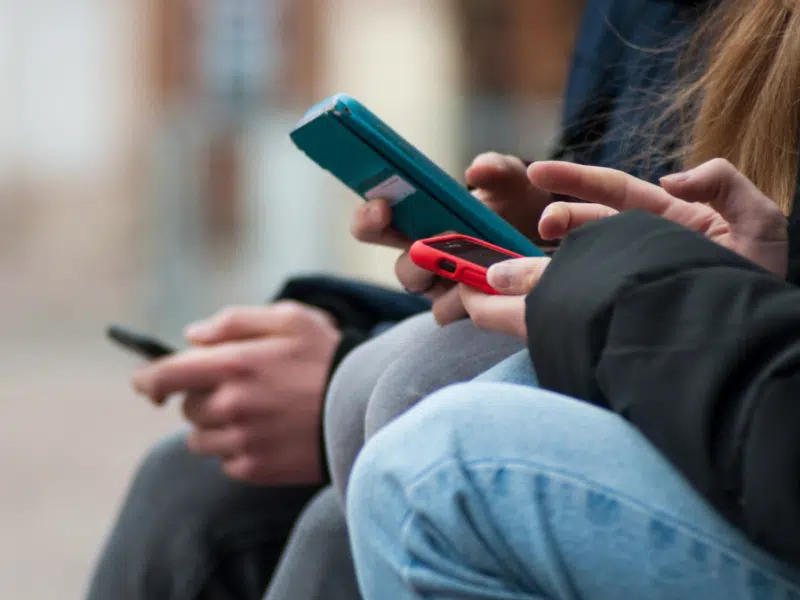 Two white teens are on their phones. The focus of the image is on their hands interacting with the phones, with their faces and upper body not visible to the image.