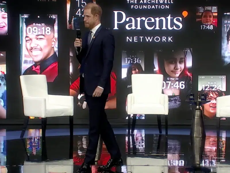 Prince Harry is walking across a stage. Holding a microphone in his right hand. He is wearing a suit. In the background you can see the text 'The Archewell Foundation' and below that and bigger 'The Parents' Network'. Around the text is screenshots of phone backgrounds of children.