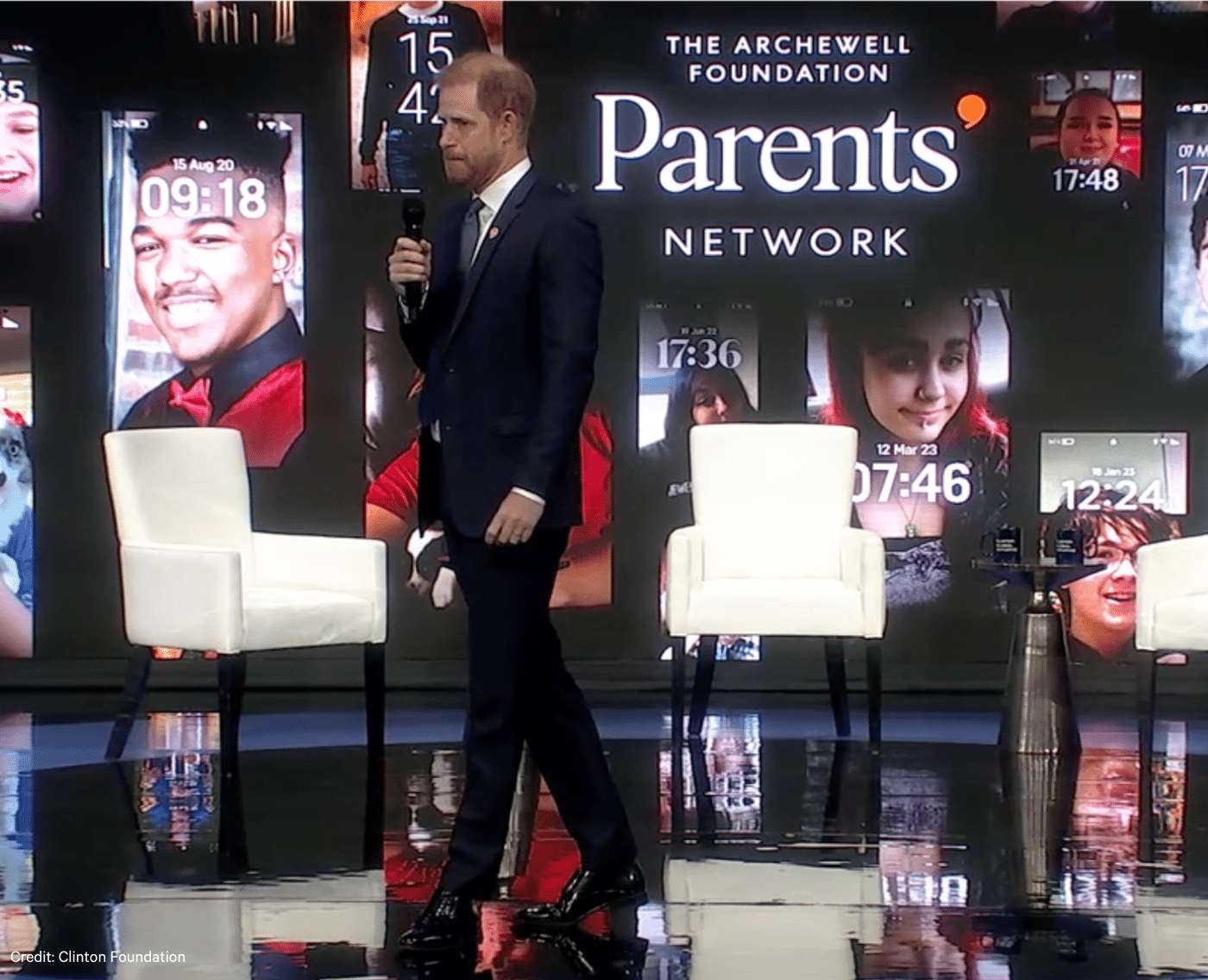 Prince Harry is walking across a stage. Holding a microphone in his right hand. He is wearing a suit. In the background you can see the text 'The Archewell Foundation' and below that and bigger 'The Parents' Network'. Around the text is screenshots of phone backgrounds of children.