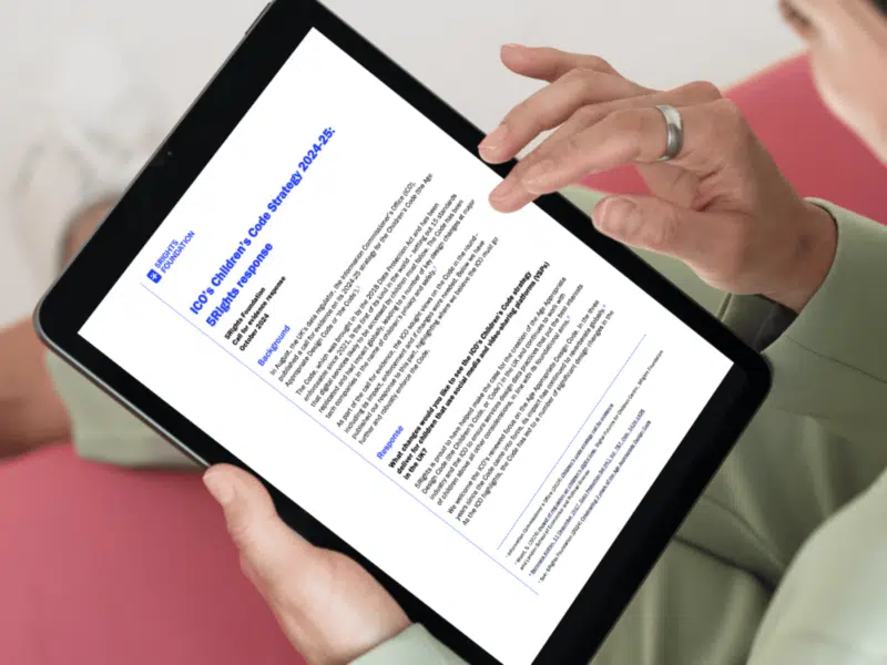 A woman is holding a tablet. She is holding the tablet in her left hand and is about to tap the screen with her right hand. On the screen is a document. She is sitting on a sofa. It is an over-the-shoulder image, and therefore, her face is only visible from the side in the top right of the image.