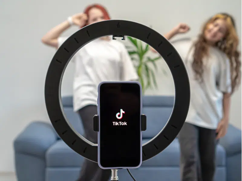 A wider shot showing two young people in the background, dancing in front of a smartphone that is set up in a ring light stand. The phone screen shows the TikTok logo, and the scene suggests that they are filming a TikTok video. The blue sofa and a potted plant are visible in the background. This is the full version of the close-up shown above.