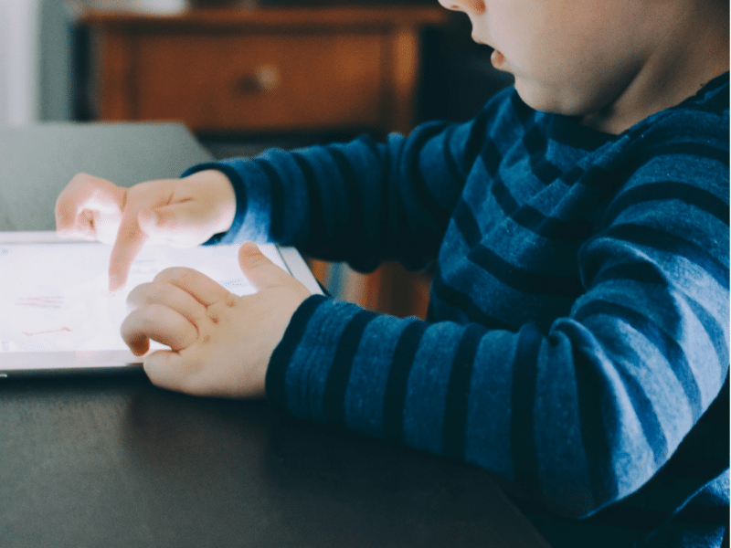 A wider shot showing the same scene in more detail. A young child, dressed in a blue and dark blue striped shirt, is sitting at a table, focused on using a tablet. The child’s fingers are touching the screen, and part of a wooden chair is visible beside them. This is the full version of the close-up shown above.