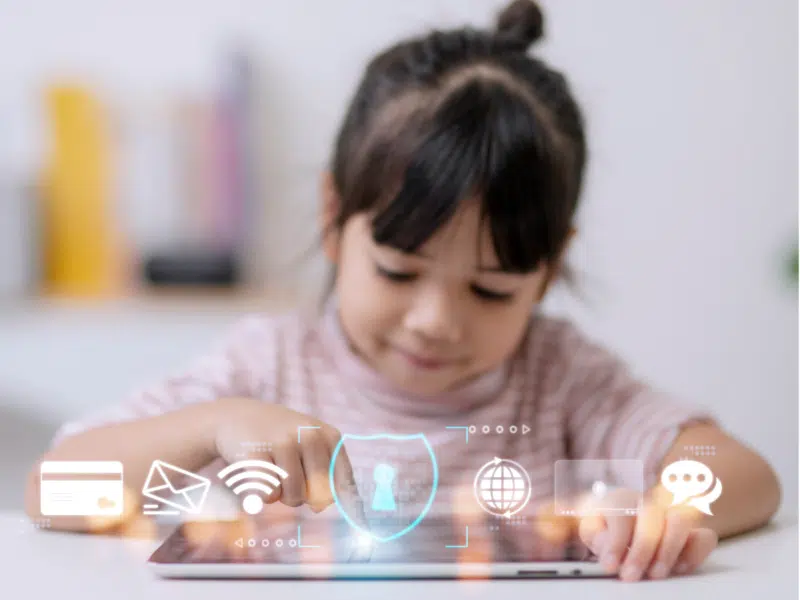 A full view of a young girl using a tablet with holographic icons hovering above the screen. The icons include a credit card, email, Wi-Fi, a shield for security, a globe, video, and a chat symbol, representing various aspects of internet usage and data protection. The background is softly blurred, drawing focus to the girl and the icons, highlighting themes of child interaction with technology and digital safety.