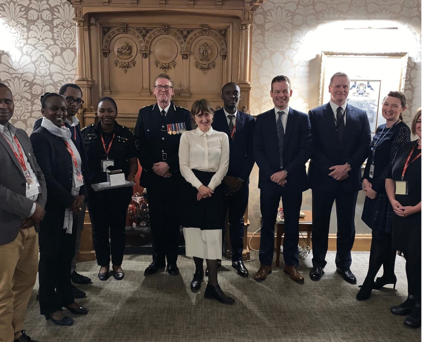 A group of people, including Rwandan Ministry representatives, Scottish Police officials, and Baroness Beeban Kidron (woman in the centre wearing a white shirt), stand together in a formal setting with an ornate wooden fireplace in the background. The group is smiling and dressed in formal attire.