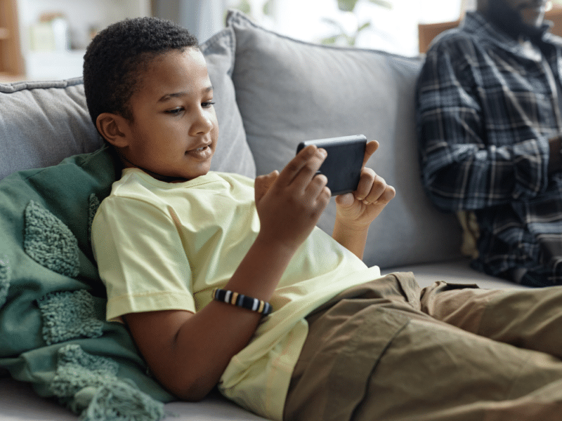 A black boy is sitting on a grey sofa, relaxed. He is wearing a yellow T-shirt and light brown cargo trousers. He is holding a smartphone horizontally and looking at it intently. You can see, to the right of the image, a black man who is out-of-focus in an open flannel shirt, looking at his own smartphone.