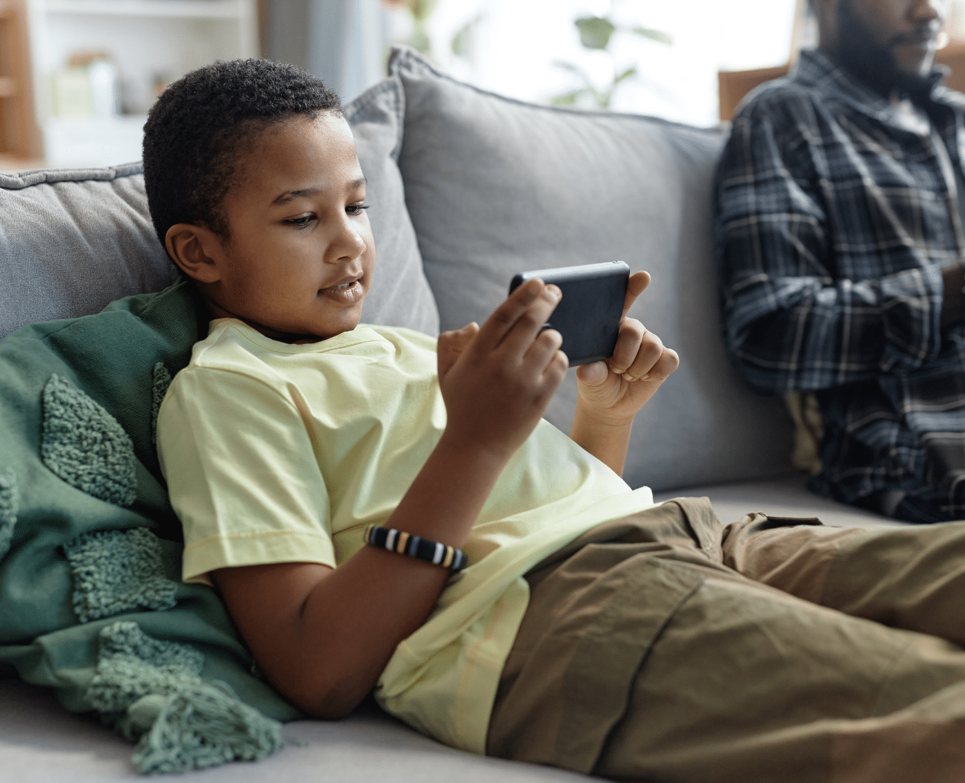 A black boy is sitting on a grey sofa, relaxed. He is wearing a yellow T-shirt and light brown cargo trousers. He is holding a smartphone horizontally and looking at it intently. You can see, to the right of the image, a black man who is out-of-focus in an open flannel shirt, looking at his own smartphone. 