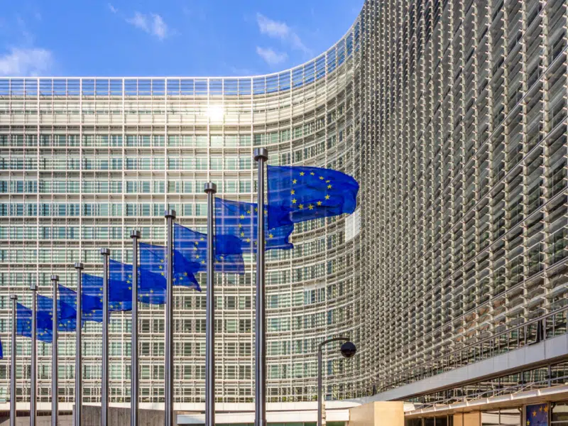 A row of EU Flags in front of the European Union Commission building in Brussels. It is a bright, sunny day. In the top left of the image, you can see clear blue skies. The sun is bouncing off the glass and the metal grid that sits in front of it.