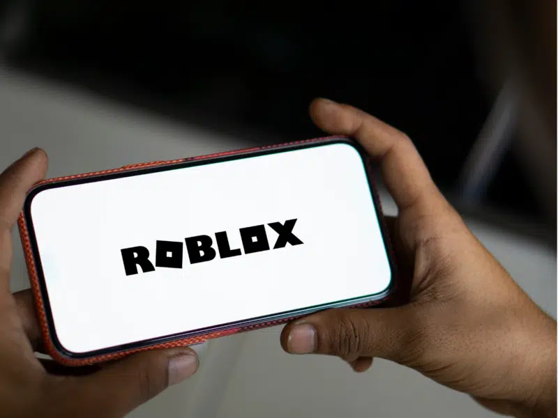 A zoomed-out view of the same scene, featuring two hands holding a smartphone with the "Roblox" logo displayed on the screen. The image provides context by showing the surroundings, emphasising the individual's engagement with the app in a more complete view.