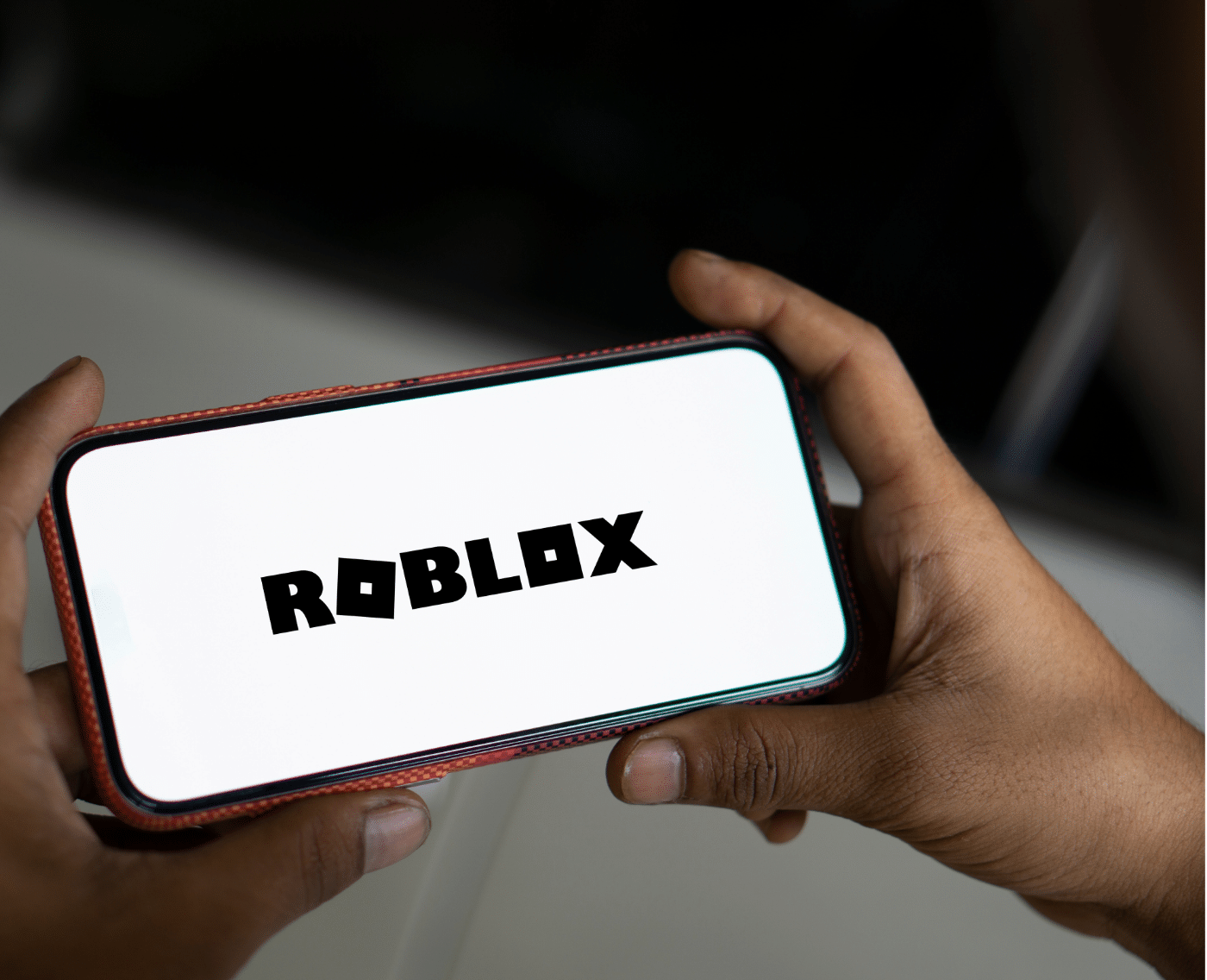 A zoomed-out view of the same scene, featuring two hands holding a smartphone with the "Roblox" logo displayed on the screen. The image provides context by showing the surroundings, emphasising the individual's engagement with the app in a more complete view.