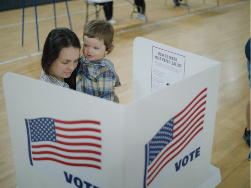 A wider view of a voting scene showing a mother holding her young child in her arms as they stand behind a voting booth marked with the American flag and "VOTE." The surrounding area of the voting hall is visible, with other individuals in the background preparing to vote.