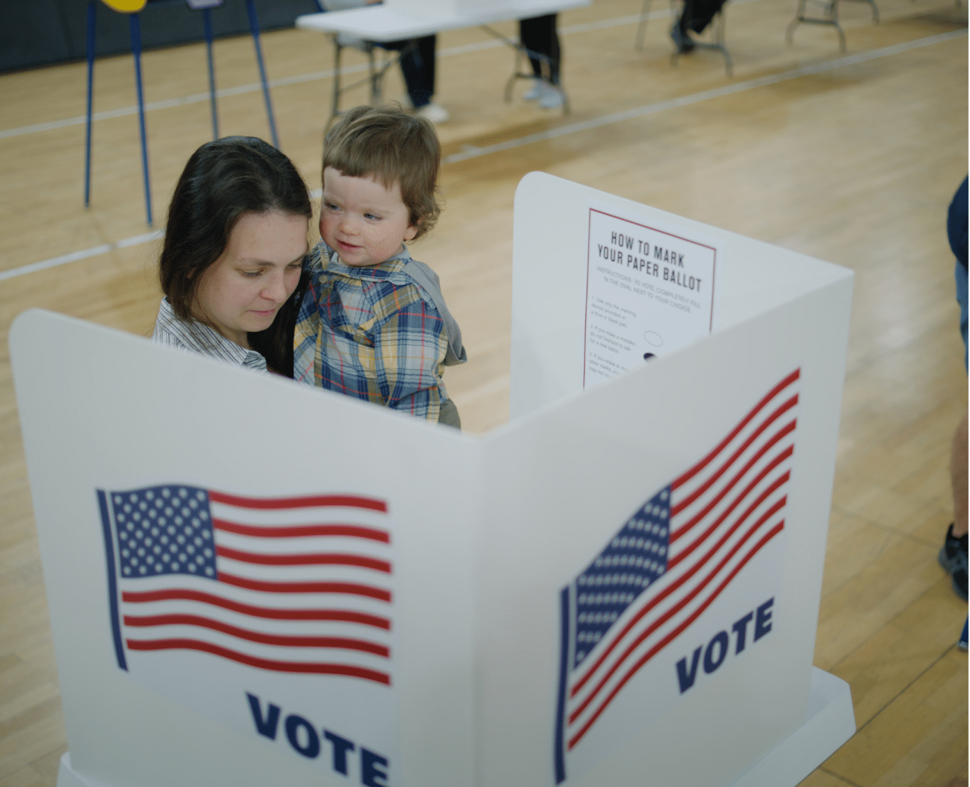 A wider view of a voting scene showing a mother holding her young child in her arms as they stand behind a voting booth marked with the American flag and "VOTE." The surrounding area of the voting hall is visible, with other individuals in the background preparing to vote.