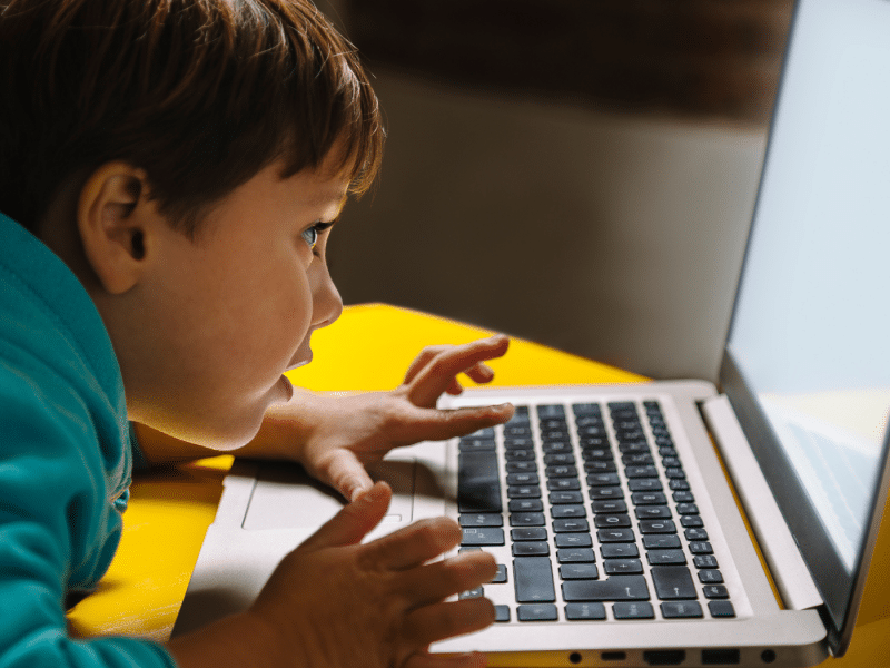 A close-up of a little boy in a teal hoodie on a laptop. He is lent over a yellow desk and has his left thumb resting on the trackpad, seemingly navigating the laptop. He is wide-eyed and very engaged.