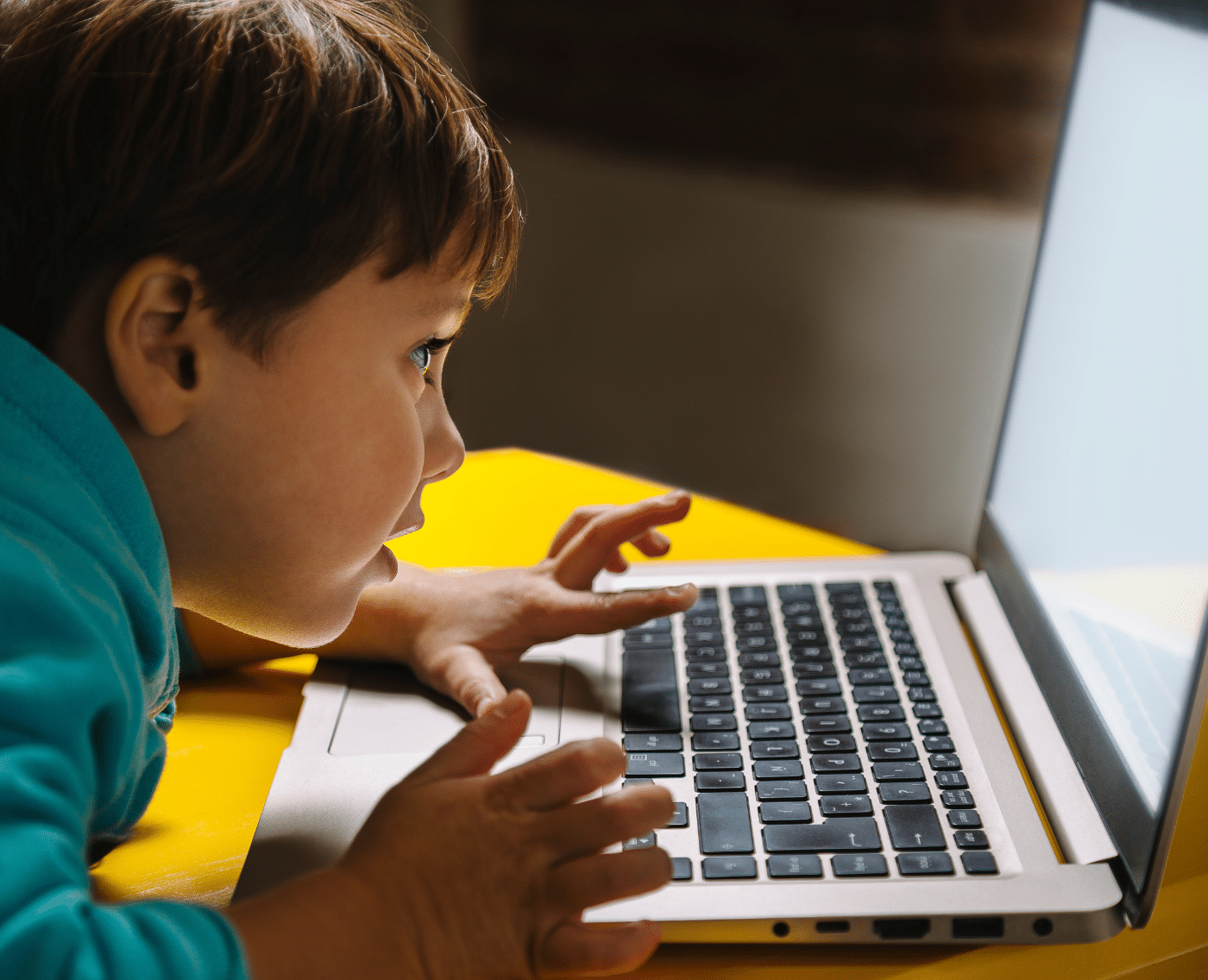 A close-up of a little boy in a teal hoodie on a laptop. He is lent over a yellow desk and has his left thumb resting on the trackpad, seemingly navigating the laptop. He is wide-eyed and very engaged.