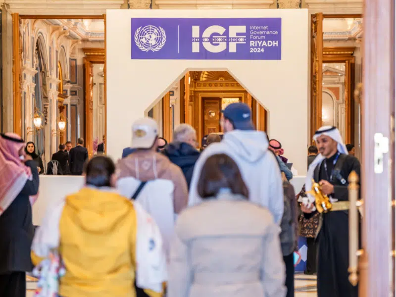 A wider view of the Internet Governance Forum 2024 entrance in Riyadh, capturing the sign as well as a bustling crowd entering the venue. The image features a mix of participants, including men in traditional Middle Eastern attire and visitors in casual and formal clothing, set against the backdrop of an elaborately decorated hall with luxurious architectural details.