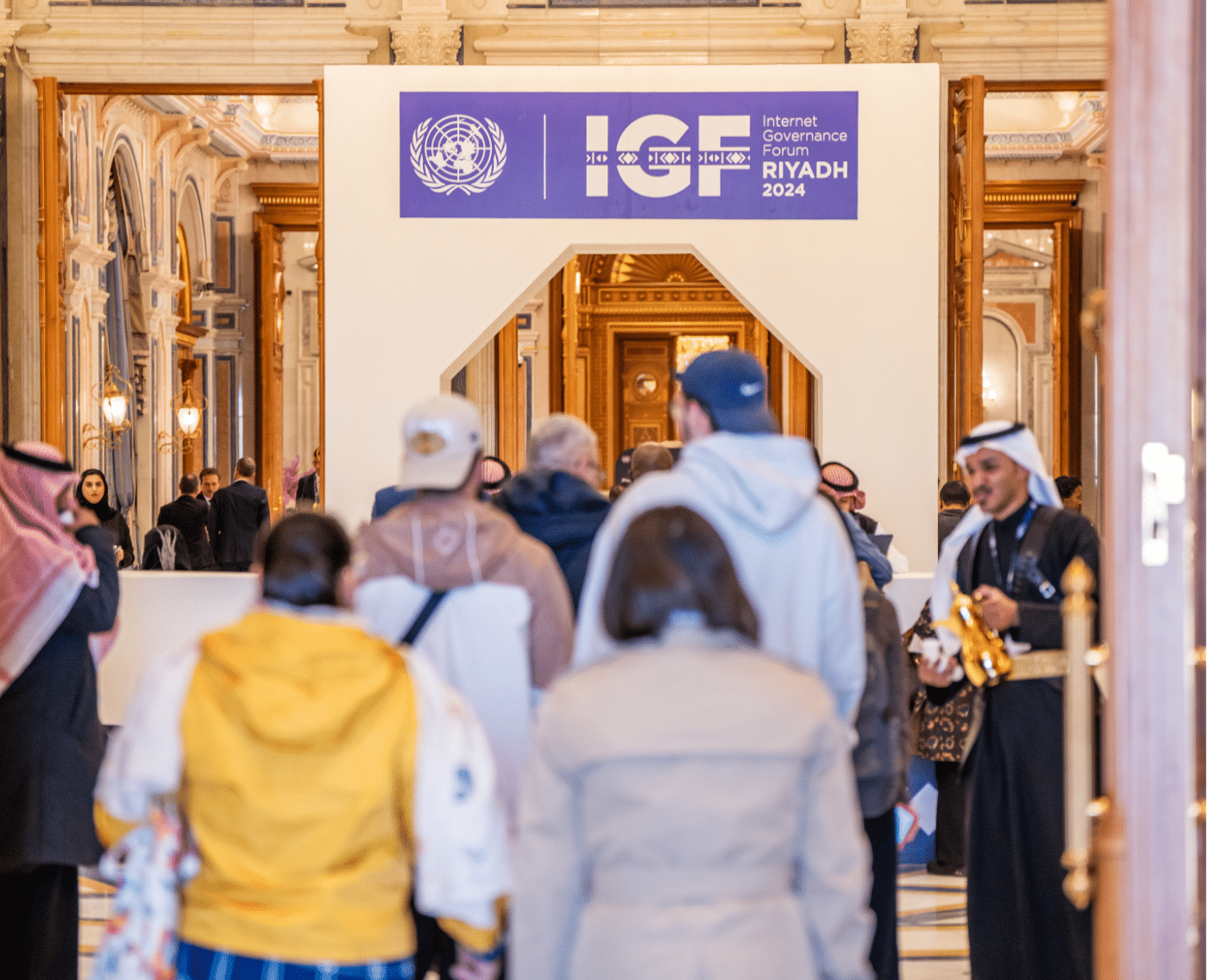 A wider view of the Internet Governance Forum 2024 entrance in Riyadh, capturing the sign as well as a bustling crowd entering the venue. The image features a mix of participants, including men in traditional Middle Eastern attire and visitors in casual and formal clothing, set against the backdrop of an elaborately decorated hall with luxurious architectural details.