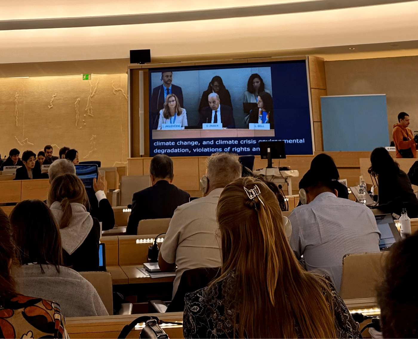 A room full of professionally-dressed people sat in a row of circled seating. With a monitor displaying the attendee speaking.