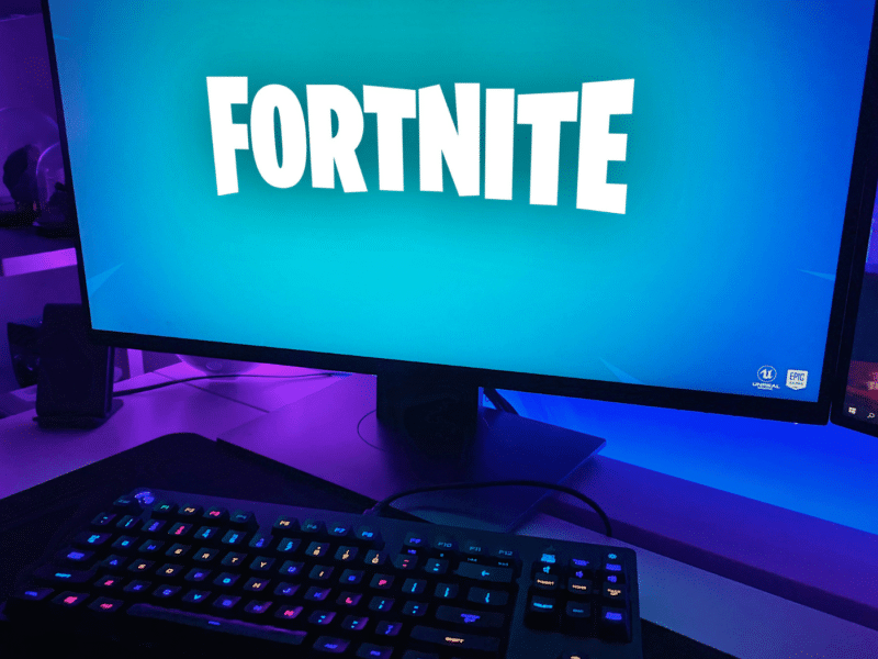 A close-up of a monitor with 'Fortnite' displayed across it, in the game's recognisable white font of all caps in a playful, comic style, with a baby blue background. In the foreground of the image is a black keyboard with a colourful backlit keyboard.