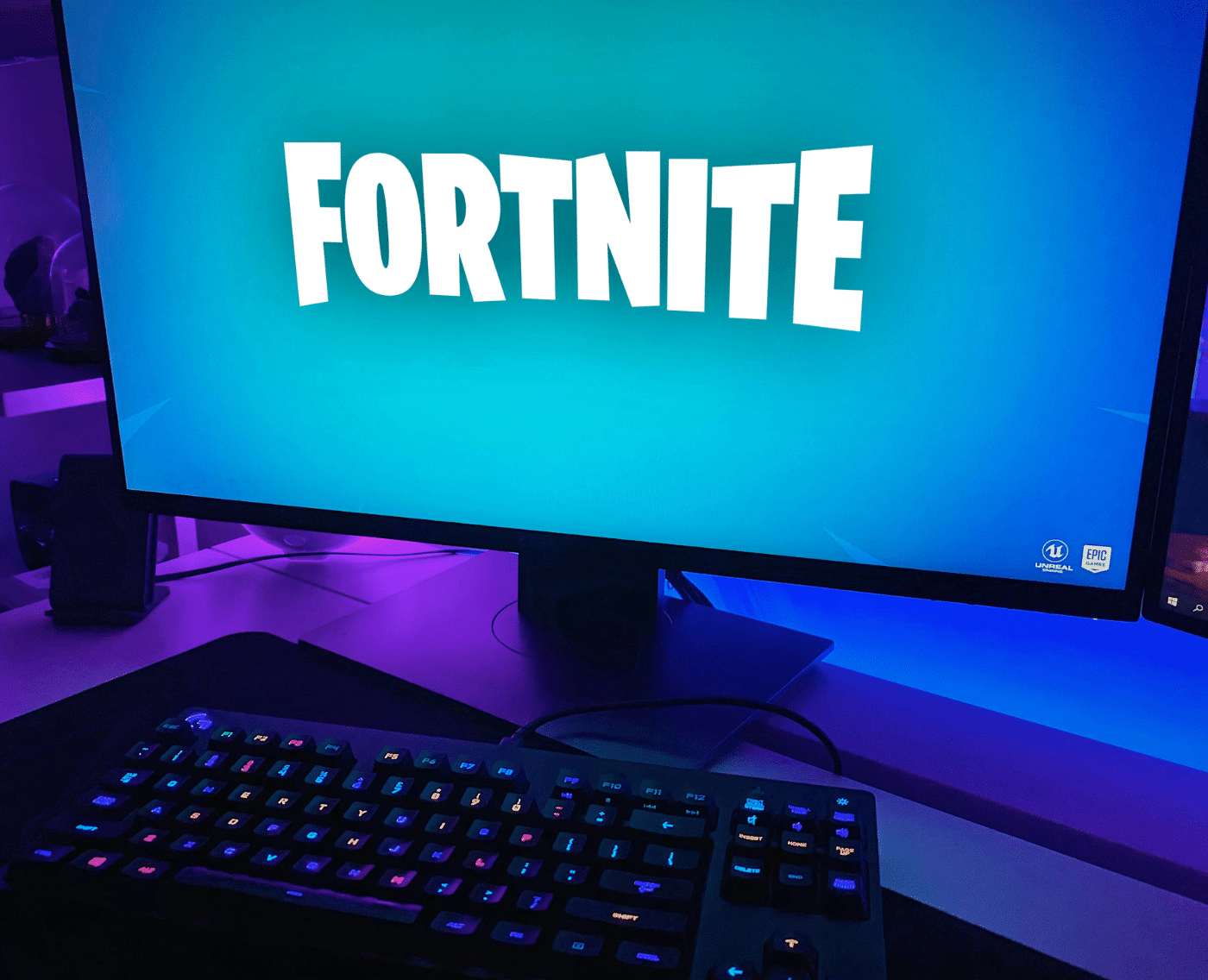 A close-up of a monitor with 'Fortnite' displayed across it, in the game's recognisable white font of all caps in a playful, comic style, with a baby blue background. In the foreground of the image is a black keyboard with a colourful backlit keyboard.