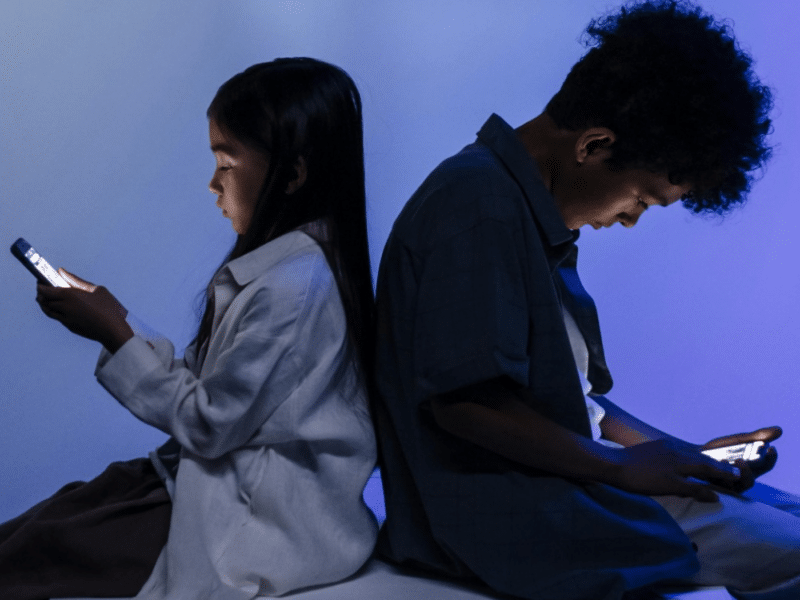 A boy and a girl are both sitting on a white box, back-to-back, both looking at their respective smartphones. The girl, a young Asian girl, is wearing a white overshirt and sits upright. The boy, seemingly a few years old, has tight curls and is sitting slouched with his phone, in both hands, rested on his thighs. The light from their phones is bouncing off their faces. The white backdrop appears lit by a purple light.