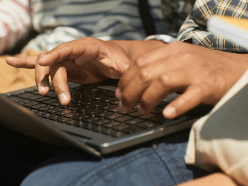 A close-up of a black child typing on a laptop. You can only see their hands typing on the keyboard, with the laptop resting on their lap. They appear to be sitting next to others.