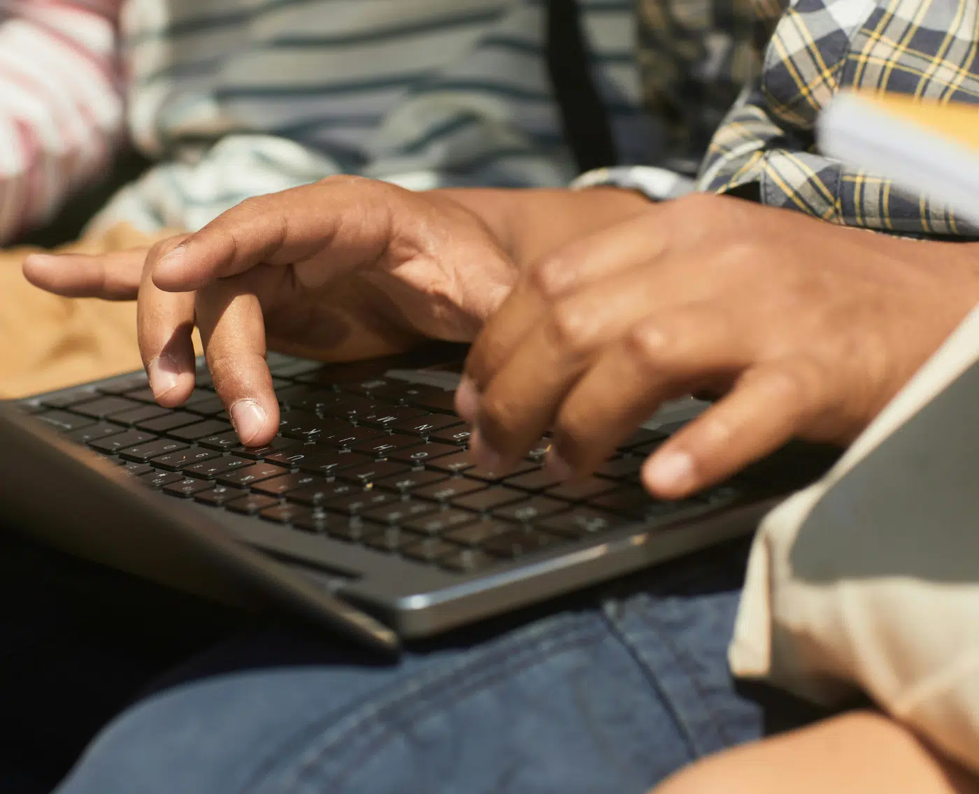 A close-up of a black child typing on a laptop. You can only see their hands typing on the keyboard, with the laptop resting on their lap. They appear to be sitting next to others.