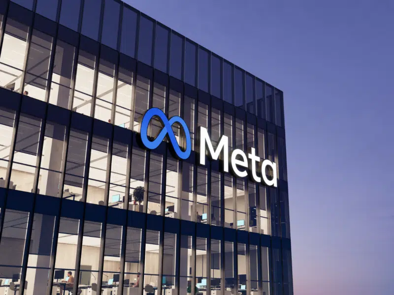 An image of a glass office building. The lights are on, and blinds open at various lengths. The image appears to have been taken in the evening, as the sky has a darkening blueish hue. Attached to the outside of the building is a blue infinity logo with the company name 'Meta', which sits across the lower middle of the top floor.