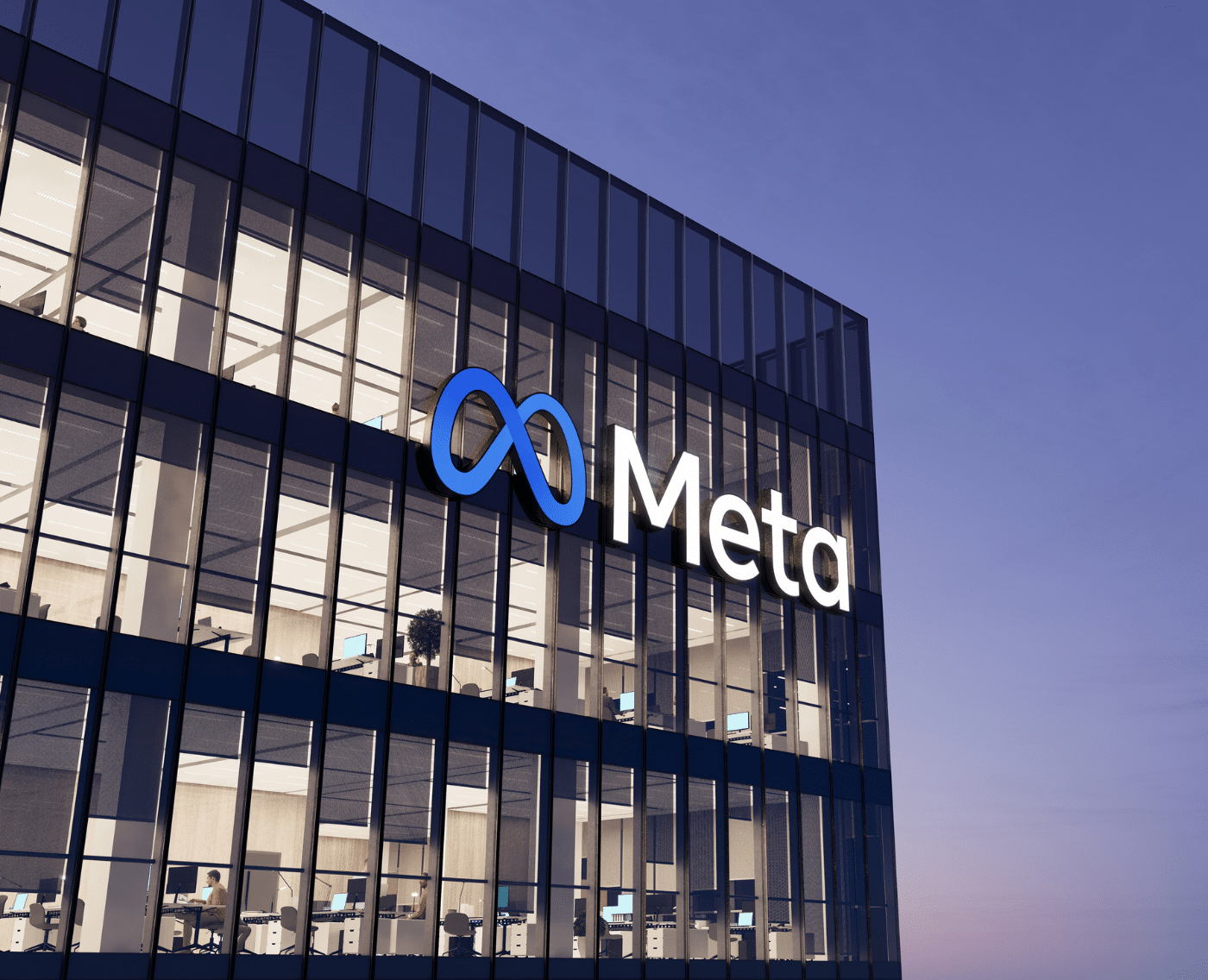 An image of a glass office building. The lights are on, and blinds open at various lengths. The image appears to have been taken in the evening, as the sky has a darkening blueish hue. Attached to the outside of the building is a blue infinity logo with the company name 'Meta', which sits across the lower middle of the top floor.