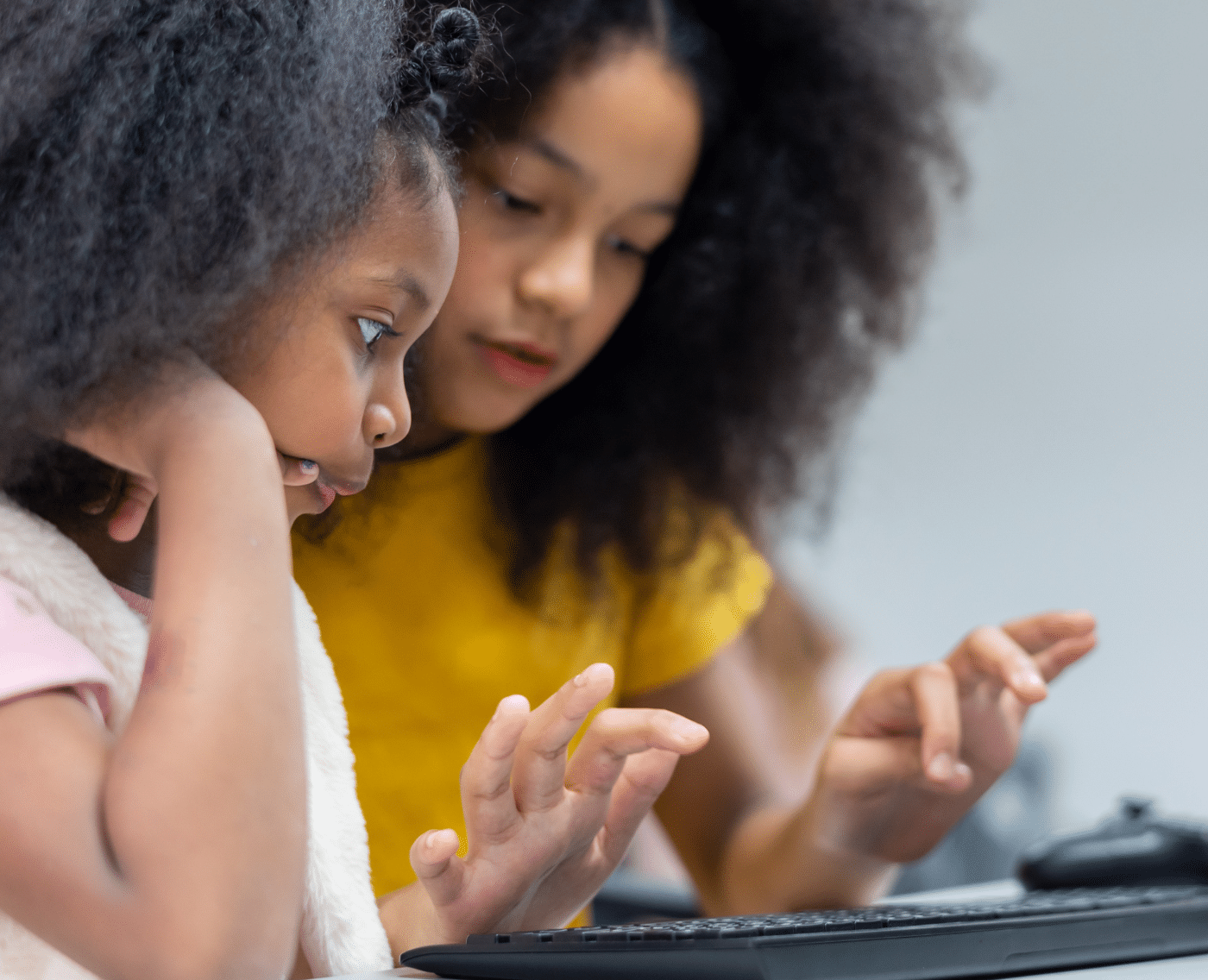 Two young black girls are on a computer. The younger of the two is in the foreground, concentrating on the older of the two typing on the keyboard.