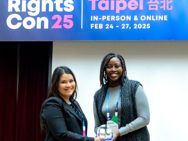 Two women smiling as one hands another an egg-shaped clear glass with a royal blue squared-glass base award. They are visibly on a stage with a Rights Con 25 banner across the top of the stage.