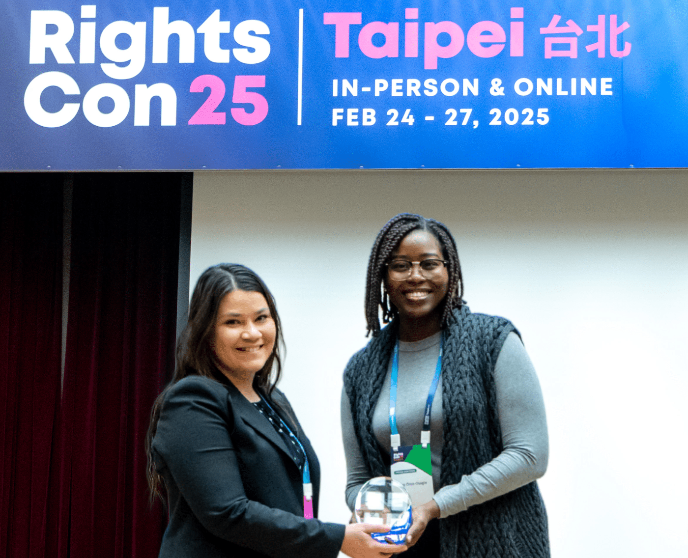 Two women smiling as one hands another an egg-shaped clear glass with a royal blue squared-glass base award. They are visibly on a stage with a Rights Con 25 banner across the top of the stage.