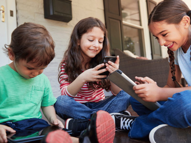 A group of children sit on the porch playing on digital devices. A young boy is on the left, playing on an iPad. A slightly older girl sits in the middle, on a phone, smiling at the screen, and the eldest, but still a young girl with plats, sits side on, cross-legged, holding and smiling at an iPad. There is a very positive mood to the photo.