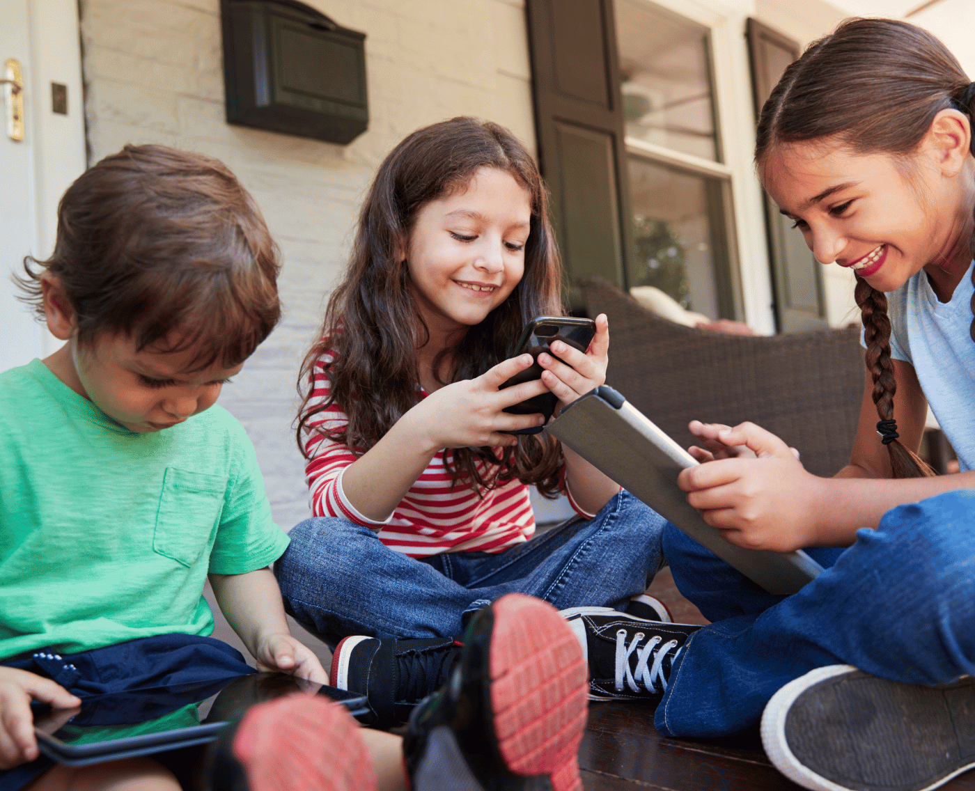 A group of children sit on the porch playing on digital devices. A young boy is on the left, playing on an iPad. A slightly older girl sits in the middle, on a phone, smiling at the screen, and the eldest, but still a young girl with plats, sits side on, cross-legged, holding and smiling at an iPad. There is a very positive mood to the photo.