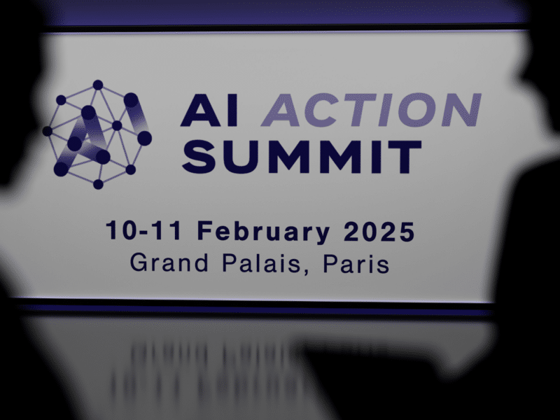 The shadows of professional-looking individuals are to the left and right of the foreground. In the middle is a projection of 'AI Action Summit' with a logo to its left, of an octagon in which all the points are connected through lines. Some lines are highlighted with a dragging effect to spell 'AI'.