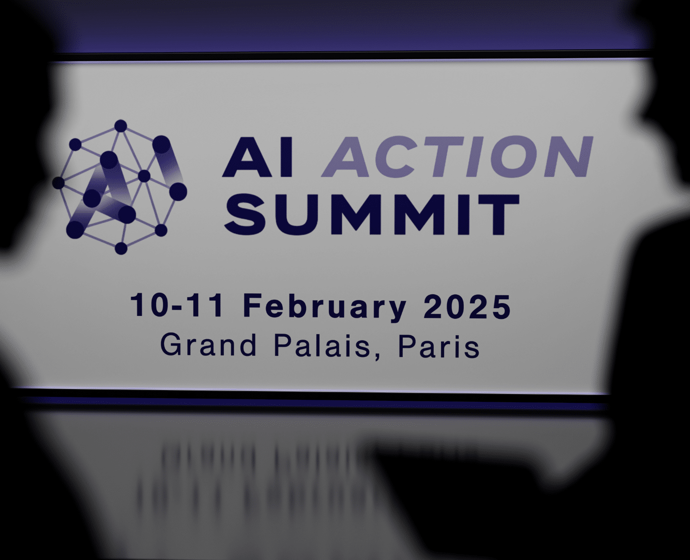 The shadows of professional-looking individuals are to the left and right of the foreground. In the middle is a projection of 'AI Action Summit' with a logo to its left, of an octagon in which all the points are connected through lines. Some lines are highlighted with a dragging effect to spell 'AI'.